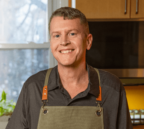 Photo of Mike, smiling in a kitchen and wearing a dark apron. 